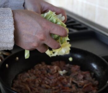 hands adding cabbage to bacon in a frying pan